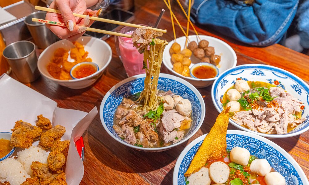 A table full of Thai food including boat noodles, skewers and fried chicken