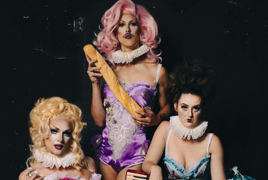 Three cabaret performers, dressed in wigs and costumes, one posed with a breadstick.