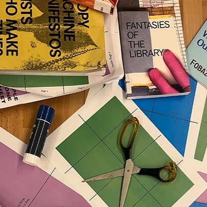 Craft paper, scissors and glue stick on a wooden table with a pile of hand-made magazines.