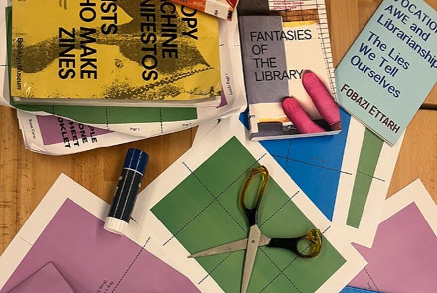 Craft paper, scissors and glue stick on a wooden table with a pile of hand-made magazines.