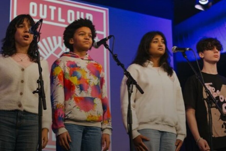 Four young students on a stage standing behind microphone stands with their arms by their sides as they recite original poetry.
