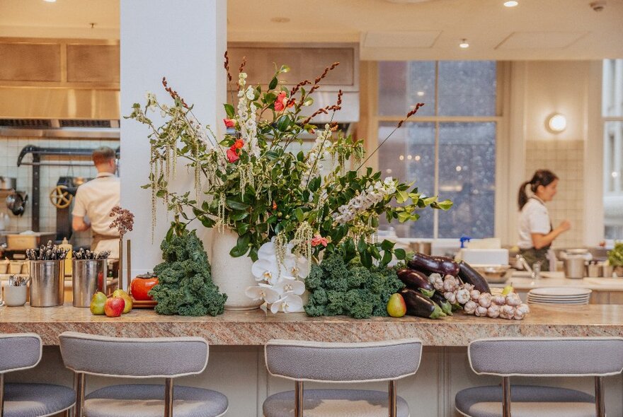 The bar counter at Hazel restaurant with piles of vegetables on display, a vase of flowers and kitchen staff in the background.