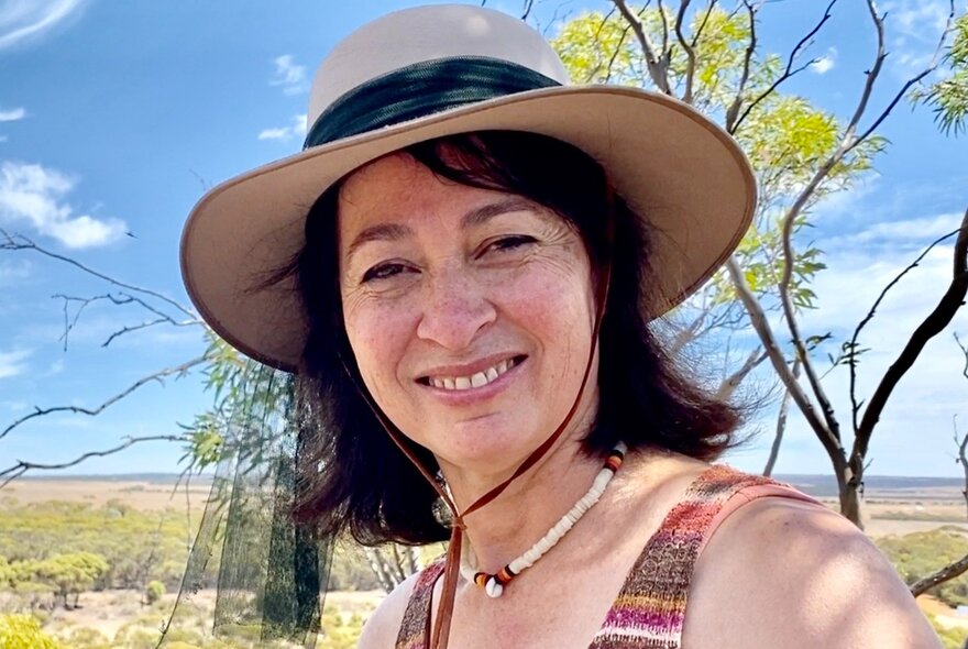 A woman taking a selfie outdoors, smiling and wearing a sunhat, a large expanse of outback scrub and a tree behind her.