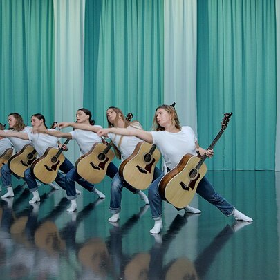 A line of woman holding guitars in an identical pose on a stage in front of a green curtain. 