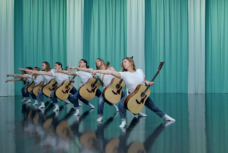 A line of woman holding guitars in an identical pose on a stage in front of a green curtain. 