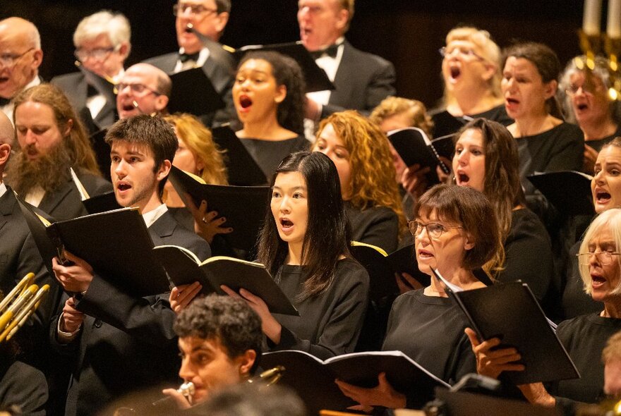 A large standing choir singing, all wearing black and holding folders of music in front of them.