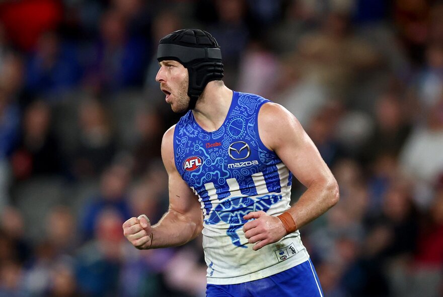 A North Melbourne AFL player celebrating on the field with both his fists clenched, he is wearing a protective head covering, there are blurred fans in the background.