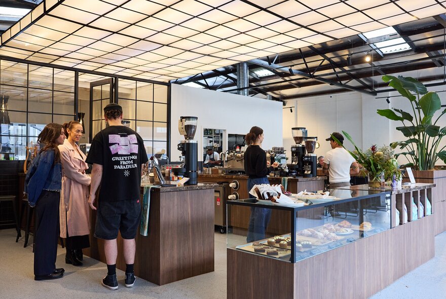 Inside a cafe with a large light fixture on the ceiling. There are three people doing a coffee workshop at the counter and two baristas making coffee.