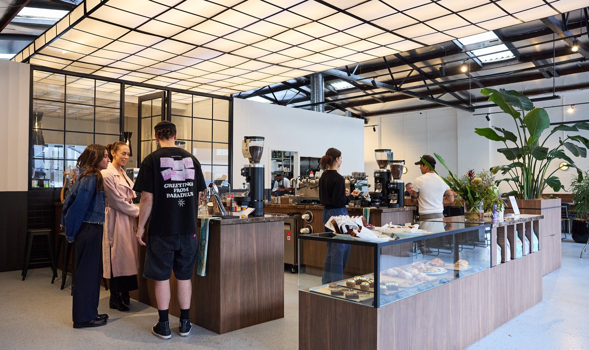 Inside a cafe with a large light fixture on the ceiling. There are three people doing a coffee workshop at the counter and two baristas making coffee.
