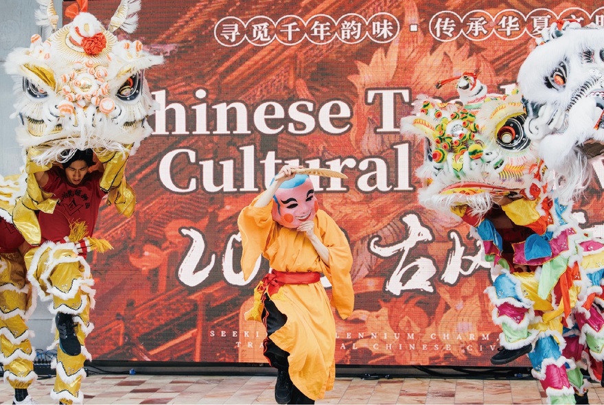 A Chinese traditional performance with a central character wearing a mask and two dragons either side, in front of a sign. 
