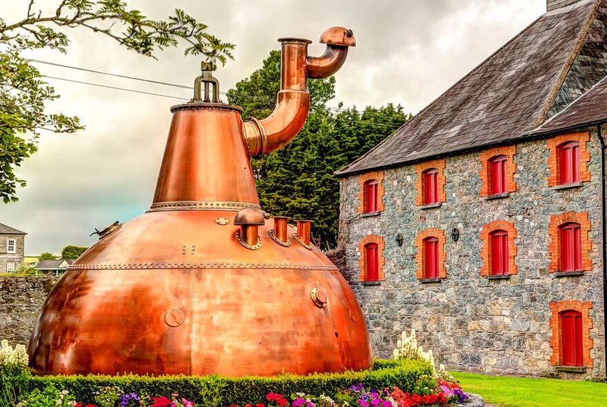A large copper pot still outdoors, the historic stone building at the Old Midleton Distillery in Ireland in the background.