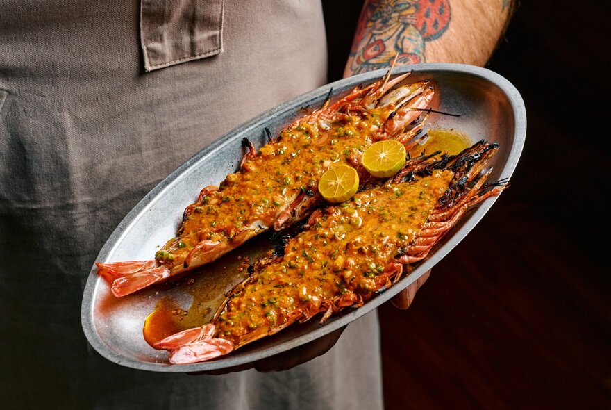A halved lobster presented on an oval silver platter with two slices of lime, held by a waiter with a tattoo on his arm.