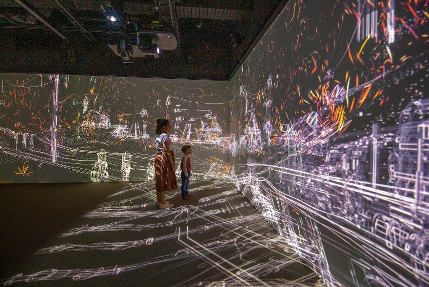 A woman and child stand watching a colourful projection across two walls.