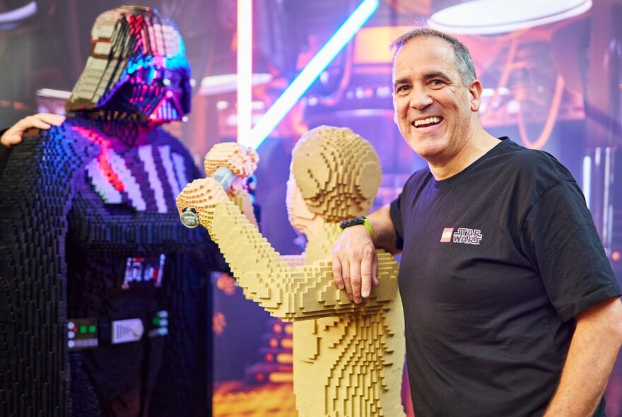 Man smiling next to life-size Darth Vader and Luke Skywalker characters made from Lego bricks.