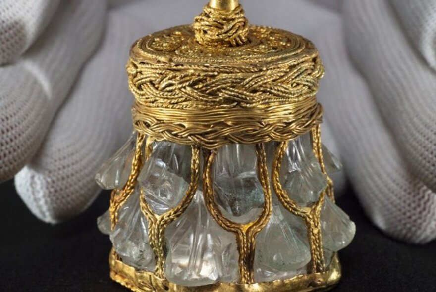 A glass and gold object from a Viking archaeological find with hands wearing white gloves behind it. 
