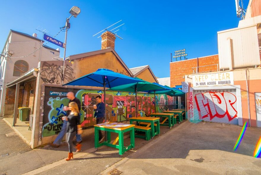 people walking past outdoor tables and umbrellas next to a street art covered building