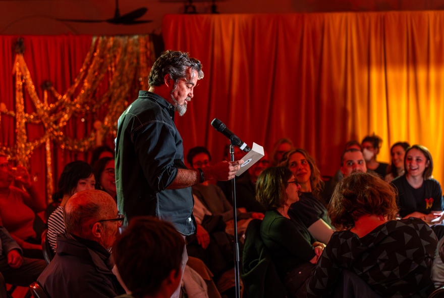 Bearded man reading from a book to a seated audience in an orange-curtained room.