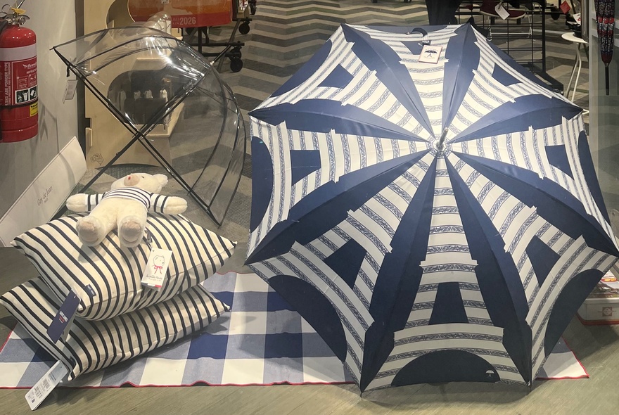 A blue and white umbrella with an Eiffel Tower pattern, displayed alongside two blue and white striped cushions, a blue and white striped teddy bear and a clear umbrella.