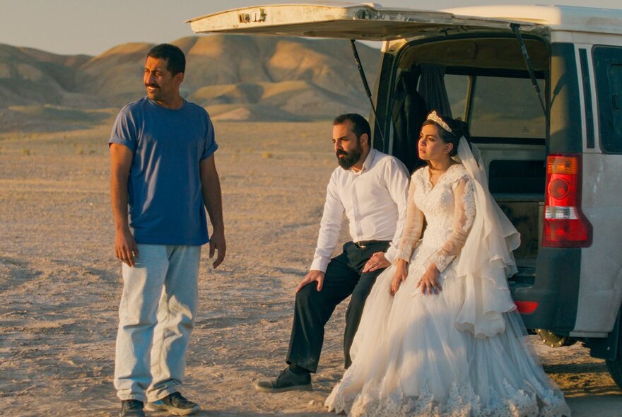 A still from an Iranian movie showing a woman in a wedding dress and her groom, sitting in the back of a van in the desert, a second man wearing jeans and a t-shirt standing nearby.