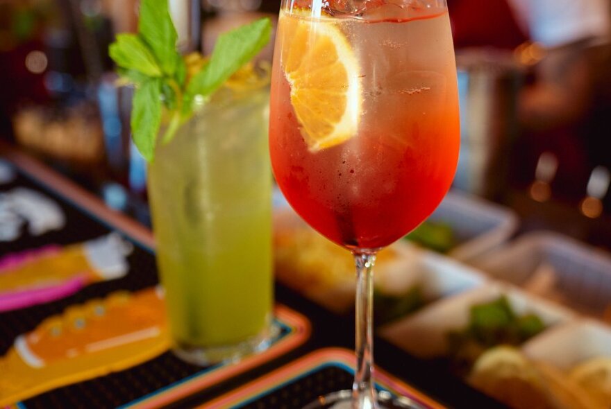 A red cocktail in a tall wine glass with a slice of orange as garnish, next to a green cocktail in a highball glass with mint leaves as garnish.