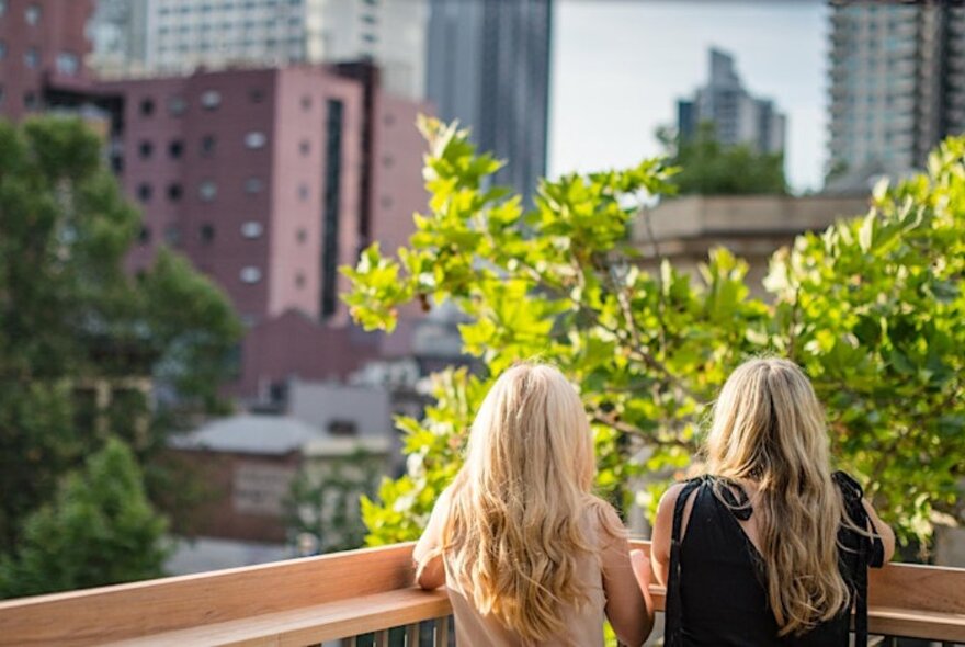 The back of two women with long blonde hair on a rooftop in the city with buildings all around. 