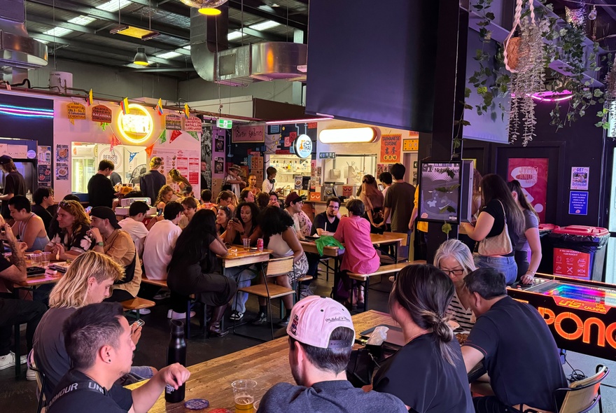 People seated at tables in a food hall, dining on a range of food and drink.