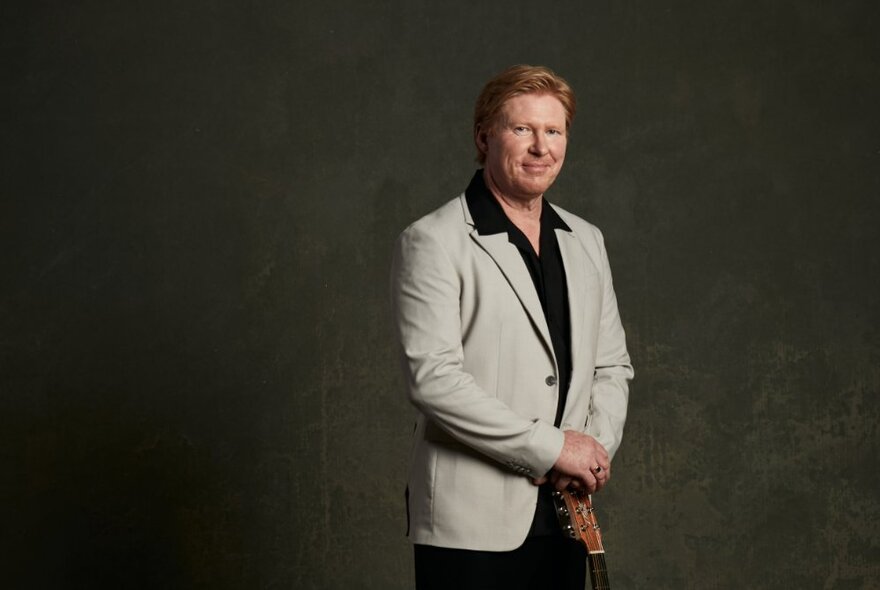 Singer Darren Coggan posing to the right of the image in a beige jacket and black shirt against a dark background. 