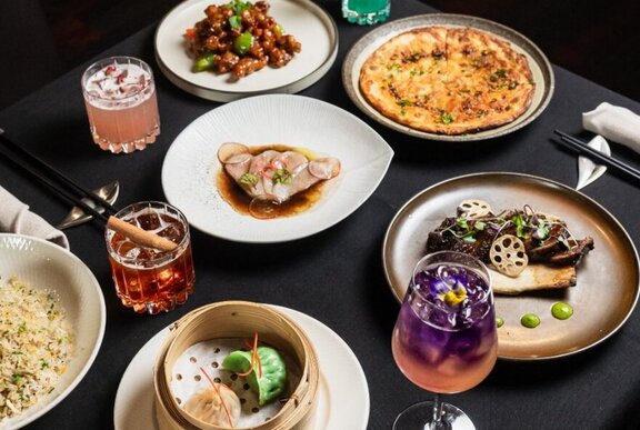 Dining table with array of Asian dishes including dumplings and noodles, with cocktails.