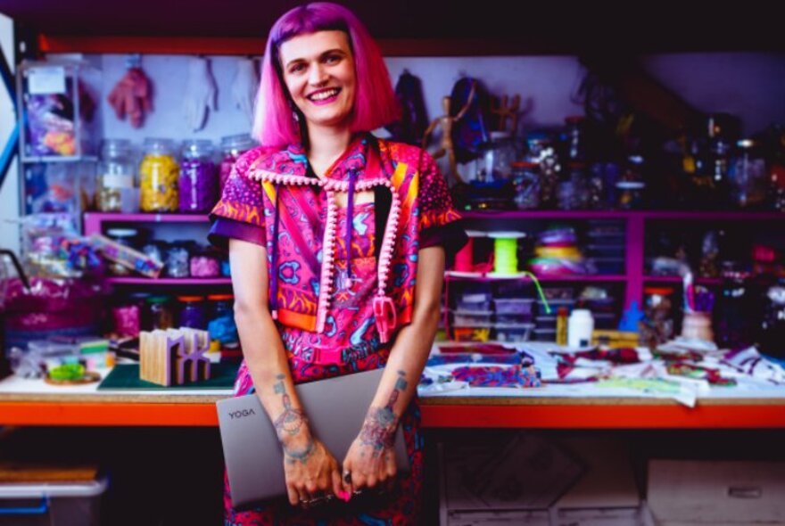 Artist Nixi Killick smiling, and holding a laptop in her hands, standing upright and leaning back against a large work table in a studio.