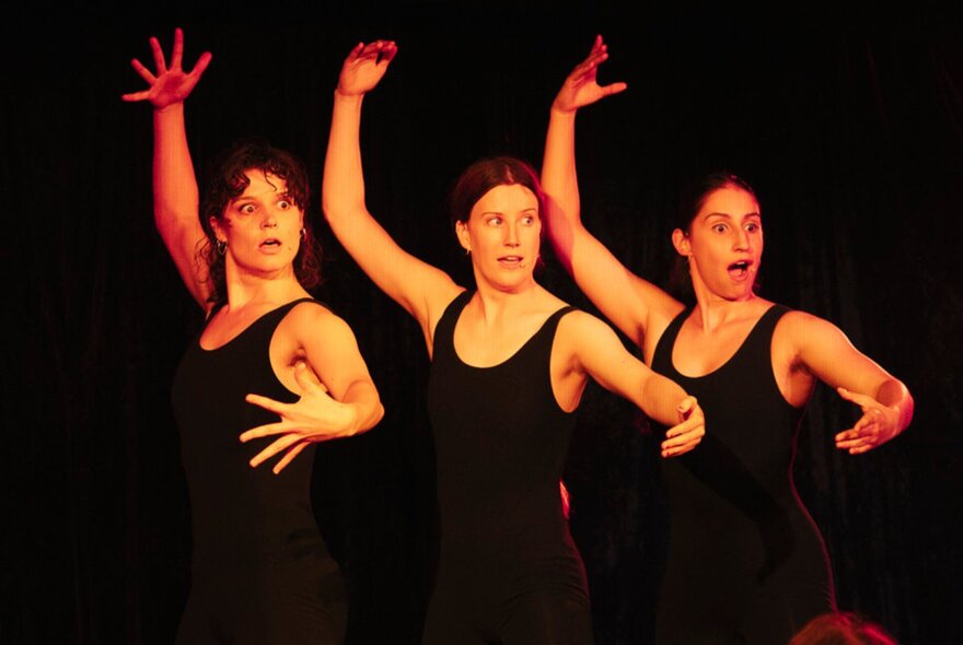 Three women in black singlets holding a ballet pose in unison with their right arms above their heads. 