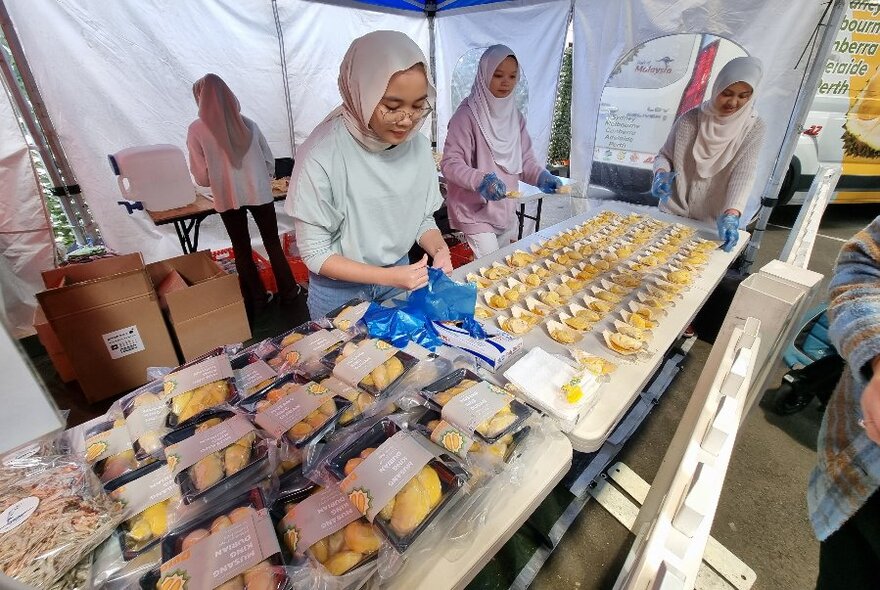 Four women behind tables of food produce, in a marquee. 