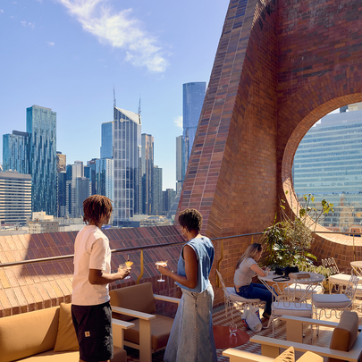 A couple on a sunny city rooftop bar with a circular window cut out of a brick wall.