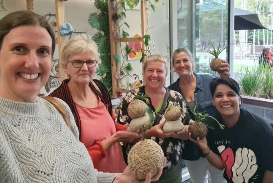 Class participants holding up their home-made kokedama balls of string topped with plants, in a workshop studio room with shelves draped with plants and crafts.