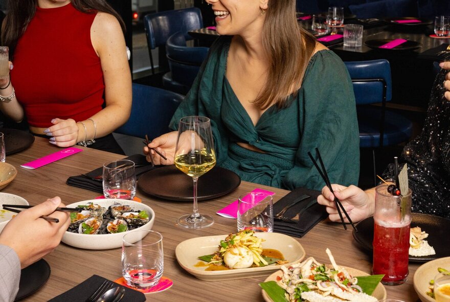 Guests at shared table with pink choptick covers, plates of food and glasses of wine.