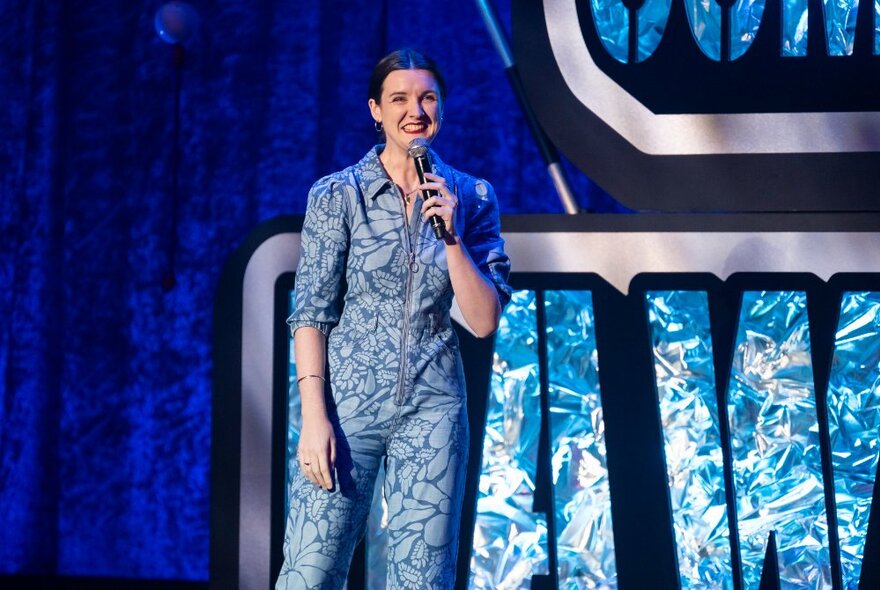 A smiling young woman standing on a stage, talking into a microphone she holds in her hand.