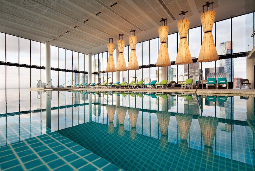 Indoor pool with infinity views outside, blue tiles and reflections of overhead lighting and poolside seating.