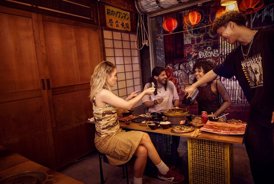Four friends sitting around a Thai barbecue grill in a small laneway restaurant with glowing lanterns hanging from the ceiling.