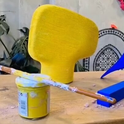 A yellow painted object shaped like a  mushroom sits on a wooden table alongside a blue bird figurine, a blue rectangular block, a yellow paint can, and a messy paintbrush balanced across them.