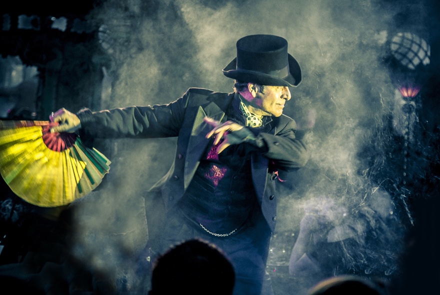 A male magician in a top hat and suit on a smoky stage, holding a yellow and green fan and performing a trick.