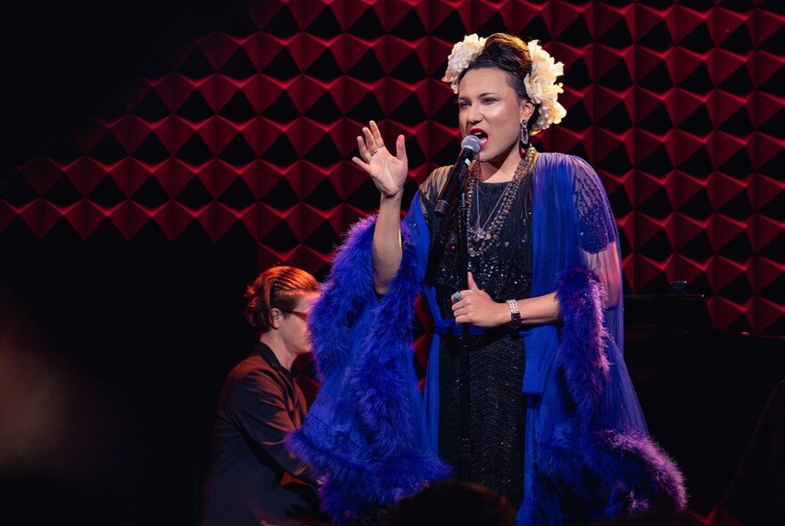 Performer singing at the microphone on a darkened stage, wearing purple robe with flowers in their hair.