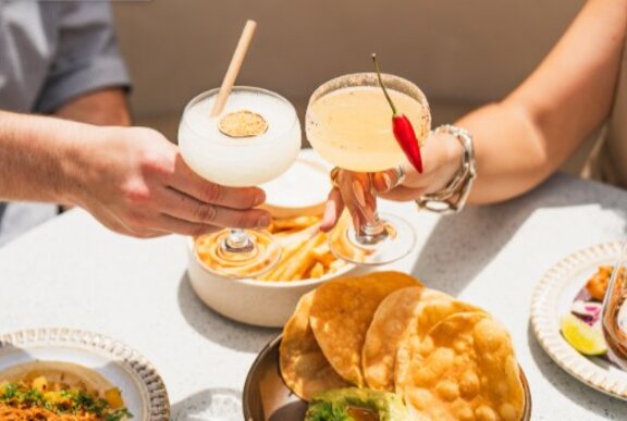 Hands clinking margaritas over a table of Mexican food.