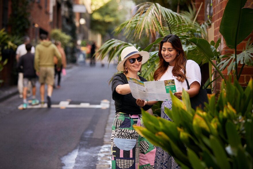 Two women looking at a map on the side of a Melbourne laneway, partially obscured by lush green plants.
