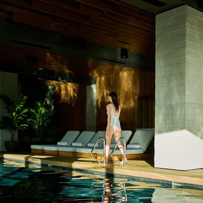 Woman walking past a hotel pool with lounge seating and dappled light.