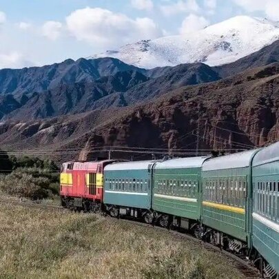 A train travelling along a curve in the track seen from outside the window of a back carriage; jagged and snow-topped mountains on the horizon behind. 