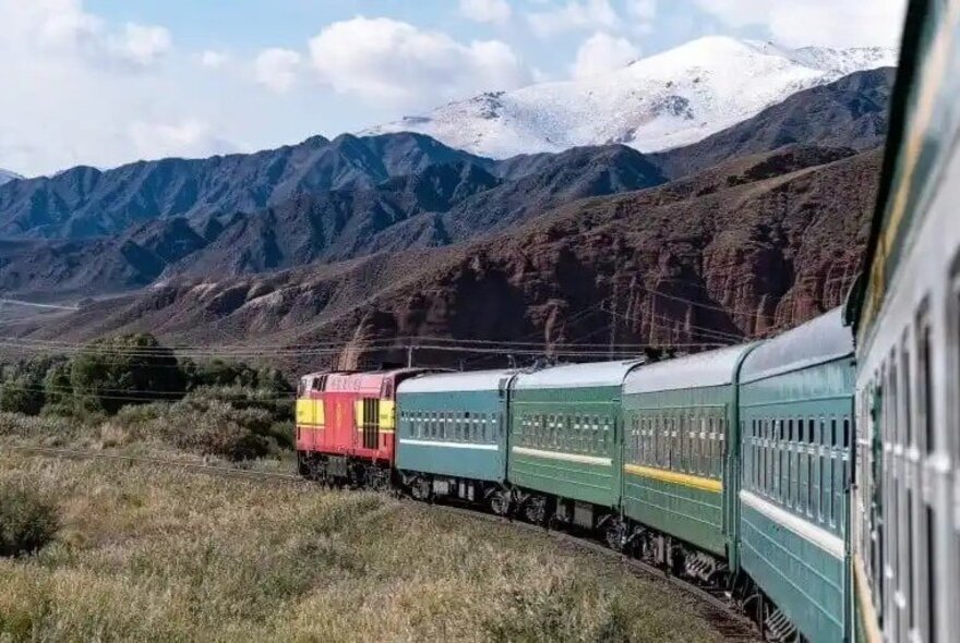 A train travelling along a curve in the track seen from outside the window of a back carriage; jagged and snow-topped mountains on the horizon behind. 