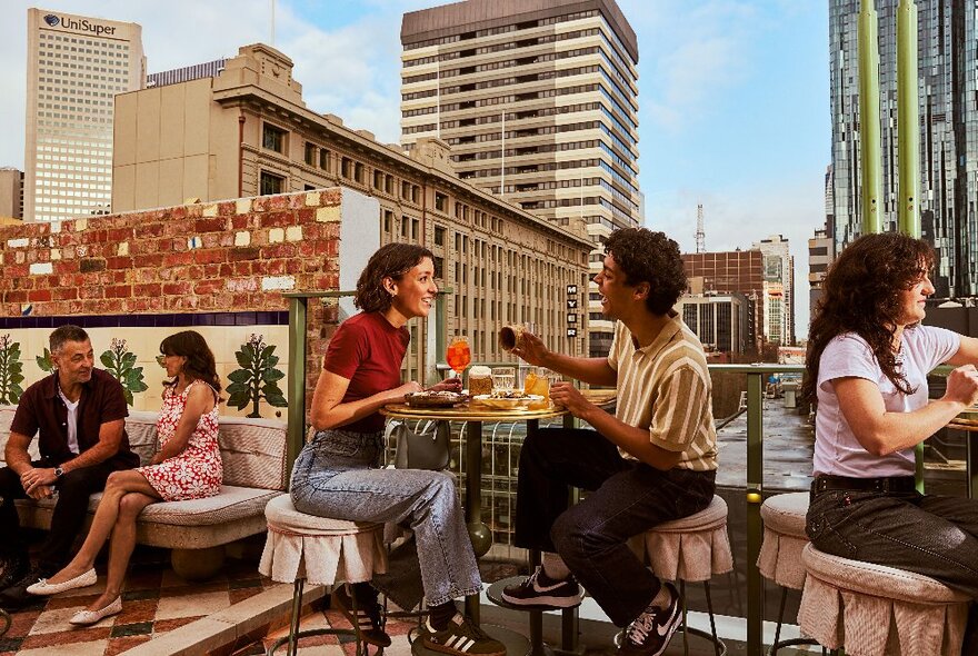 Diners at tables on an outdoor rooftop overlooking Myer and other city buildings, including central couple with drinks and snacks.