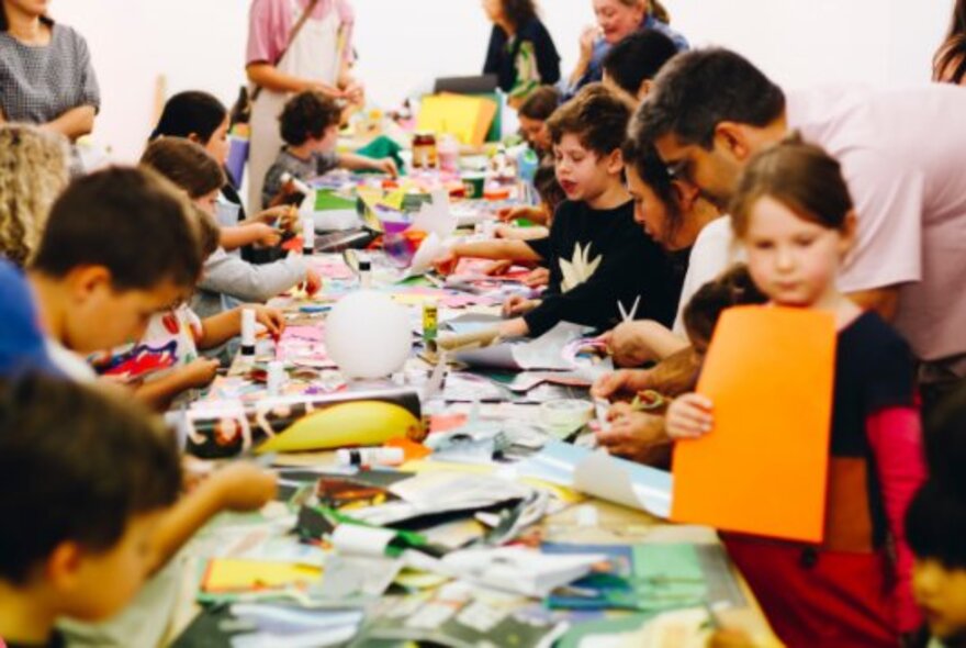 Children and adults at a long work table covered in art materials.