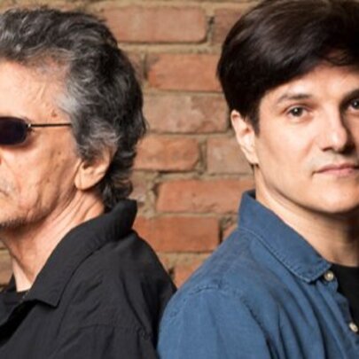 Two men standing back-to-back in front of a rustic red brick wall. 