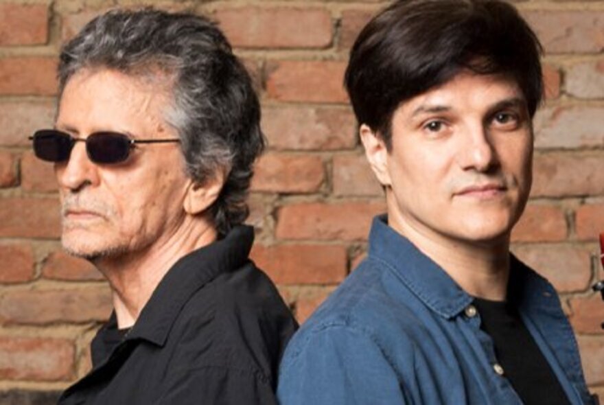 Two men standing back-to-back in front of a rustic red brick wall. 