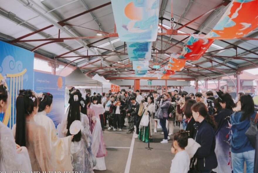 People standing in one of the sheds at Queen Victoria Market, with coloured sashes decorating the ceiling, watching a traditional dance performance.
